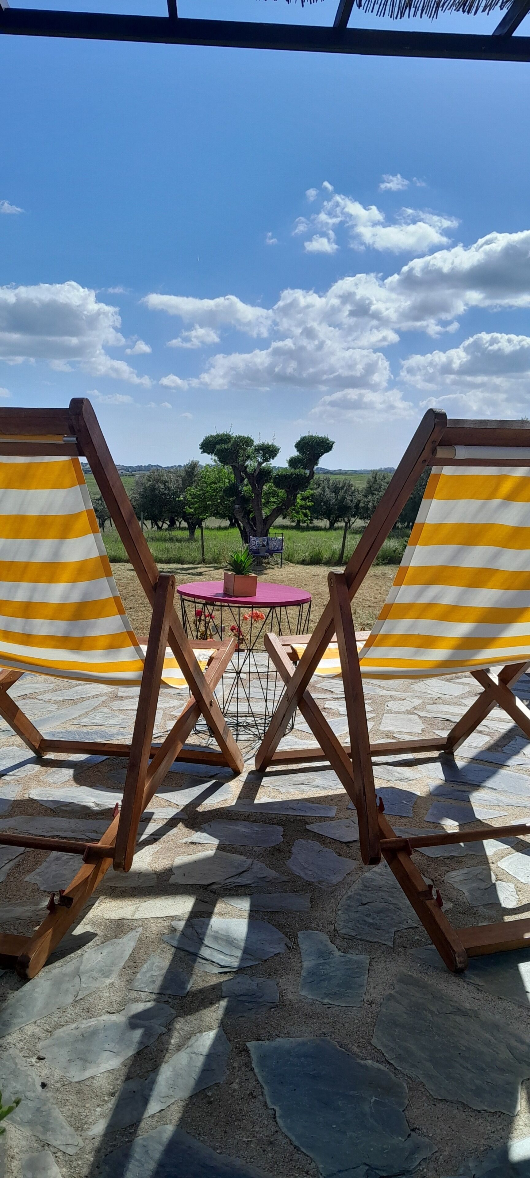 Deux fauteuils en bois, à rayures jaunes et blanches, sont placés face à un paysage verdoyant, avec des arbres et un ciel dégagé.