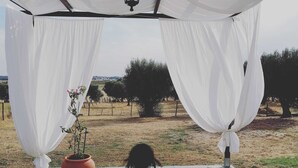 A person sitting on a stone floor under a white canopy with drapes, with a potted plant beside them. - Monte da Rua 13 - Casa de Aldeia (Redondo)