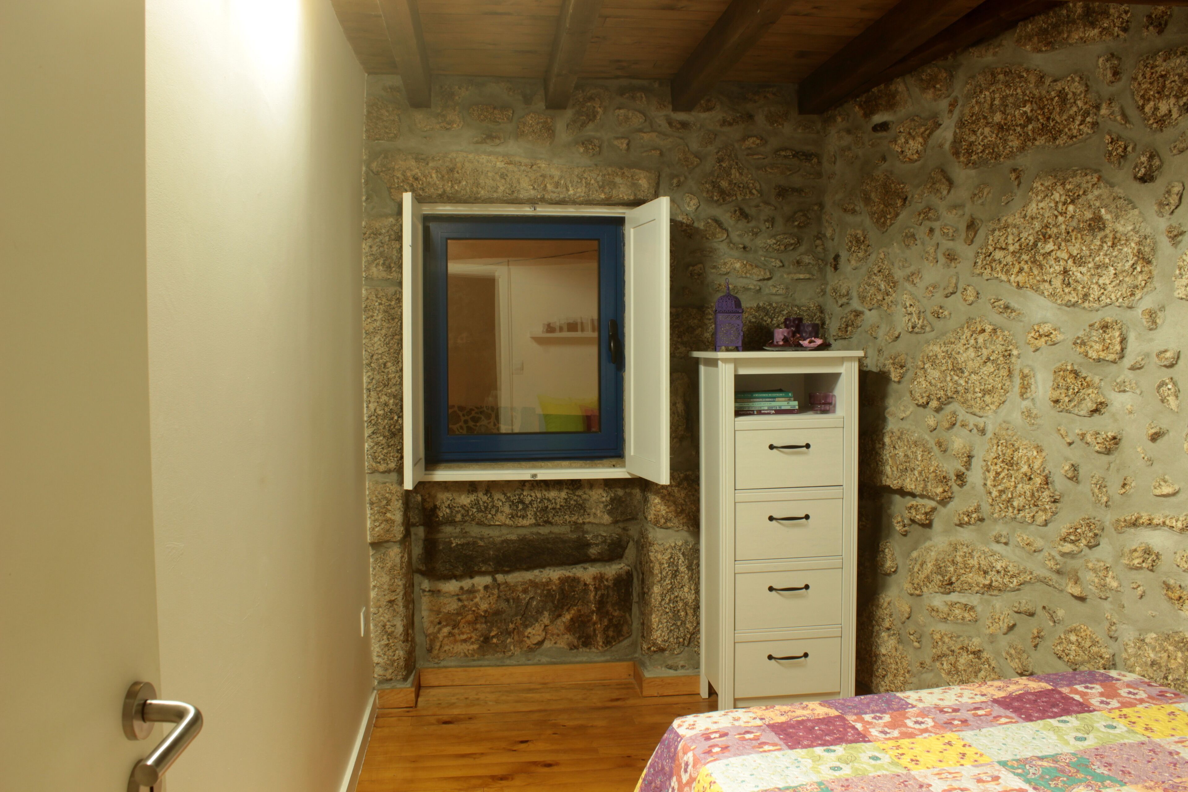 A bedroom with a stone wall, a wooden ceiling, a bed with a colorful quilt, and a white dresser with drawers.