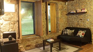 Una stanza accogliente con parete in pietra, soffitto in legno, un divano nero con cuscini, un tavolino con un libro e una finestra con persiane in legno.