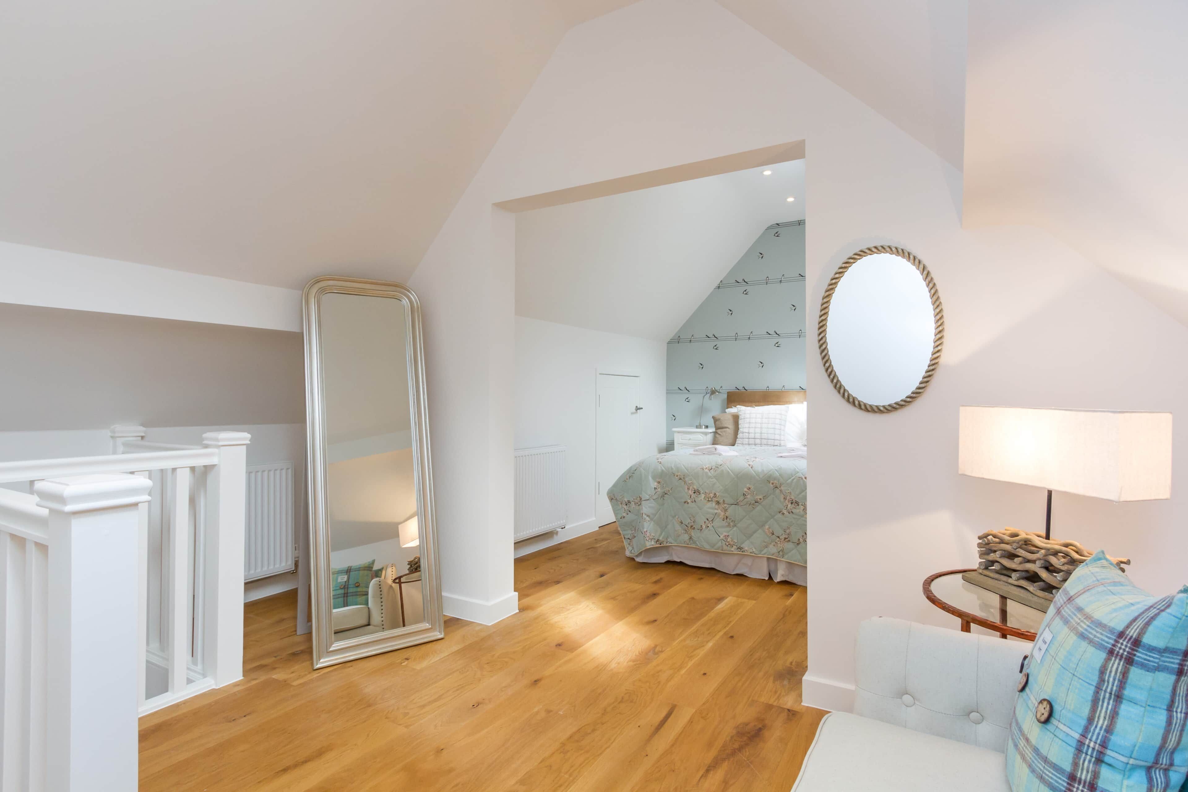 A bedroom with a large mirror, a bed with a floral duvet, a wooden floor, and a white dresser.