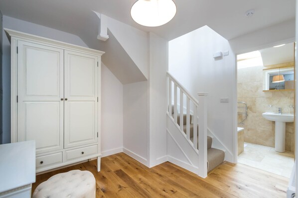 A room with a wooden floor, white walls, a white wardrobe, a staircase, and a bathroom with a sink and mirror.