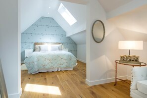 A bedroom with a sloped ceiling, a bed with floral bedding, a bedside table, a lamp, and a round mirror on the wall.