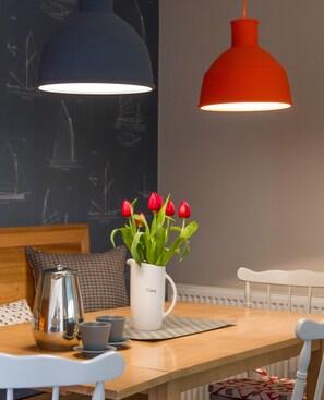 Une table à manger avec un pichet blanc, des tasses et des soucoupes grises, une théière en argent et un luminaire suspendu rouge.