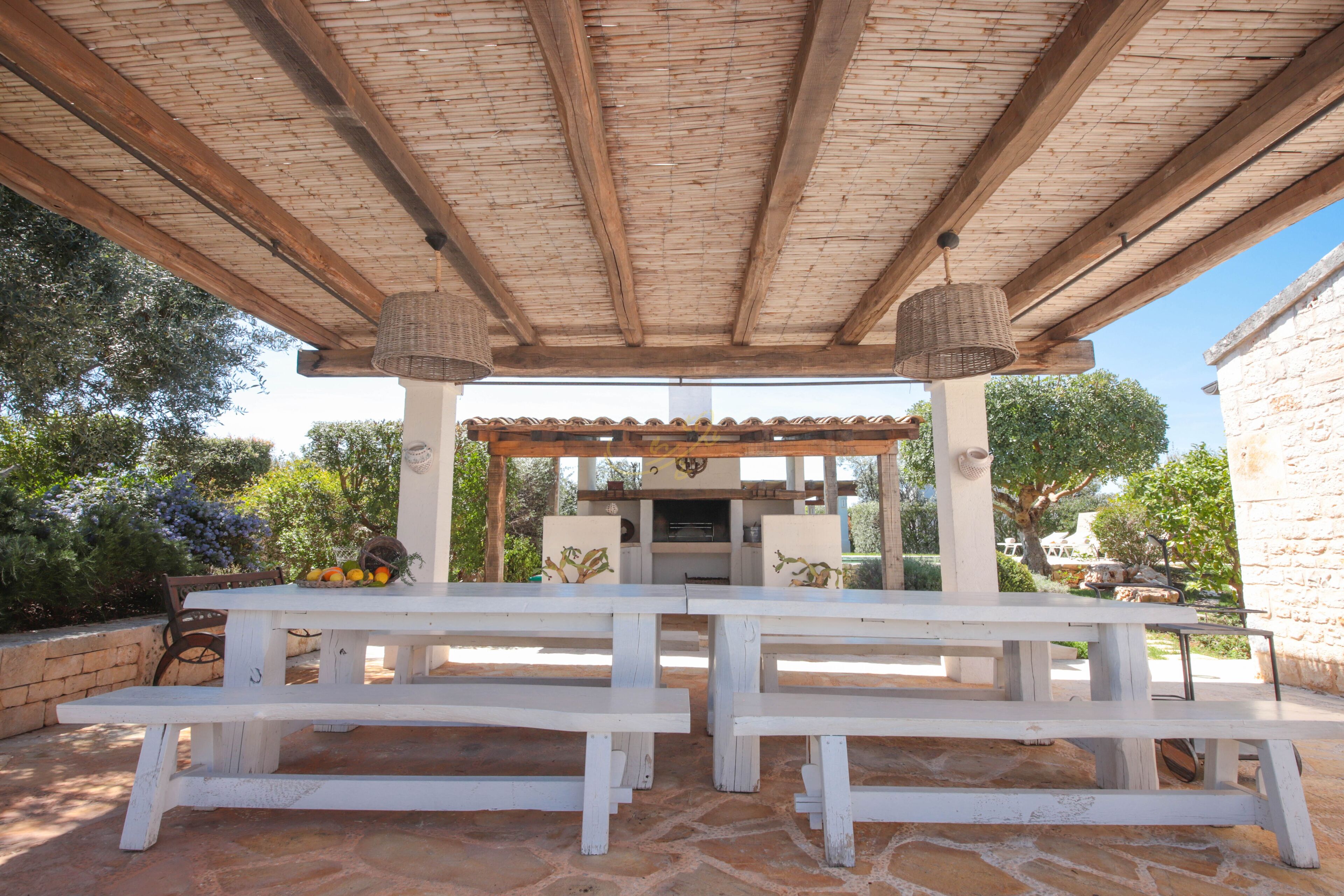 An outdoor patio with a wooden pergola, white benches, and a central grill area.