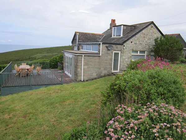 A stone house with a deck, a dining table, and a garden with pink flowers.