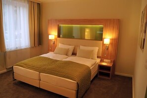 A hotel room with a double bed, two bedside tables, a nightstand, a mirror above the bed, and a window with curtains.