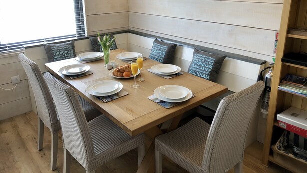 Un coin repas avec une table en bois dressée pour deux personnes, entourée de chaises en osier. On y trouve un petit vase avec des fleurs et une bouteille de jus d’orange. La pièce présente un design moderne, avec un téléviseur à écran plat fixé au mur au-dessus de la table.