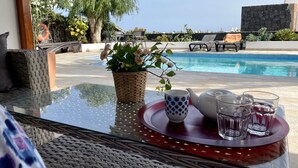 A glass table with a woven basket, a teapot, and glasses, overlooking a pool and lounge area. - Spacious Villa with Private, Sunny Garden, Sea View and Exclusive Pool  (Playa Blanca)