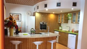A modern kitchen with a central island, bar stools, a refrigerator, an oven, a dishwasher, and wooden cabinets. - Spacious Villa with Private, Sunny Garden, Sea View and Exclusive Pool  (Playa Blanca)