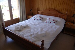 Un lit en bois avec des draps blancs et des coussins à motifs floraux, une table de chevet avec une lampe et une fenêtre avec des rideaux.