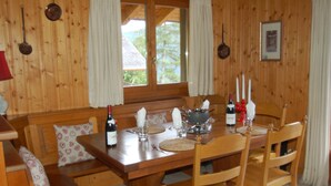 Ein Holztisch ist gedeckt mit Weingläsern, Tellern und einer Schüssel. Stühle und eine Bank mit Kissen stehen um den Tisch herum.