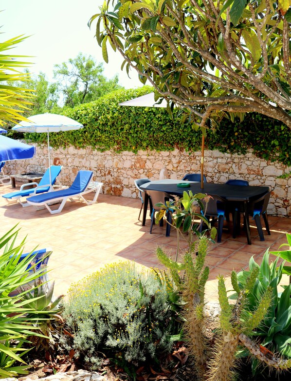 Une terrasse extérieure avec un sol en pierre, une table avec des chaises et des fauteuils bleus placés sous des parasols.