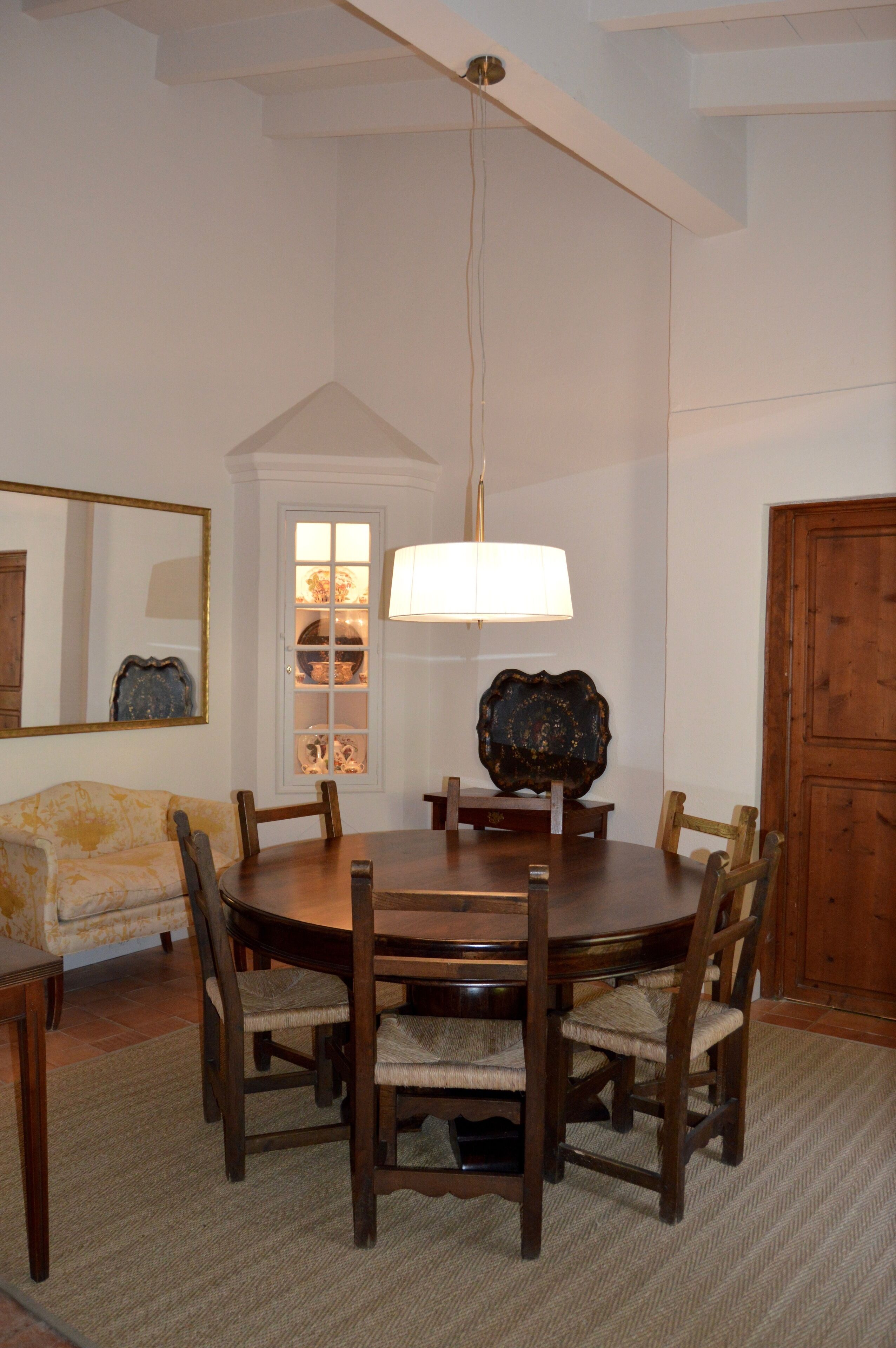 Uma sala de jantar com uma mesa redonda de madeira, seis cadeiras, um lustre, um espelho e uma porta de madeira.
