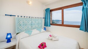 Ein Schlafzimmer mit einem Bett, einem Nachttisch, einer Lampe, einem Fenster mit Vorhängen und Blick aufs Meer.