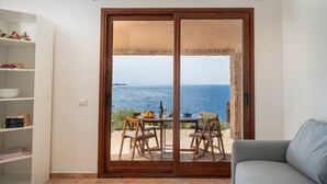 Ein Zimmer mit einer Schiebetür zum Balkon mit Meerblick. Es gibt einen runden Tisch mit Stühlen und ein Sofa.