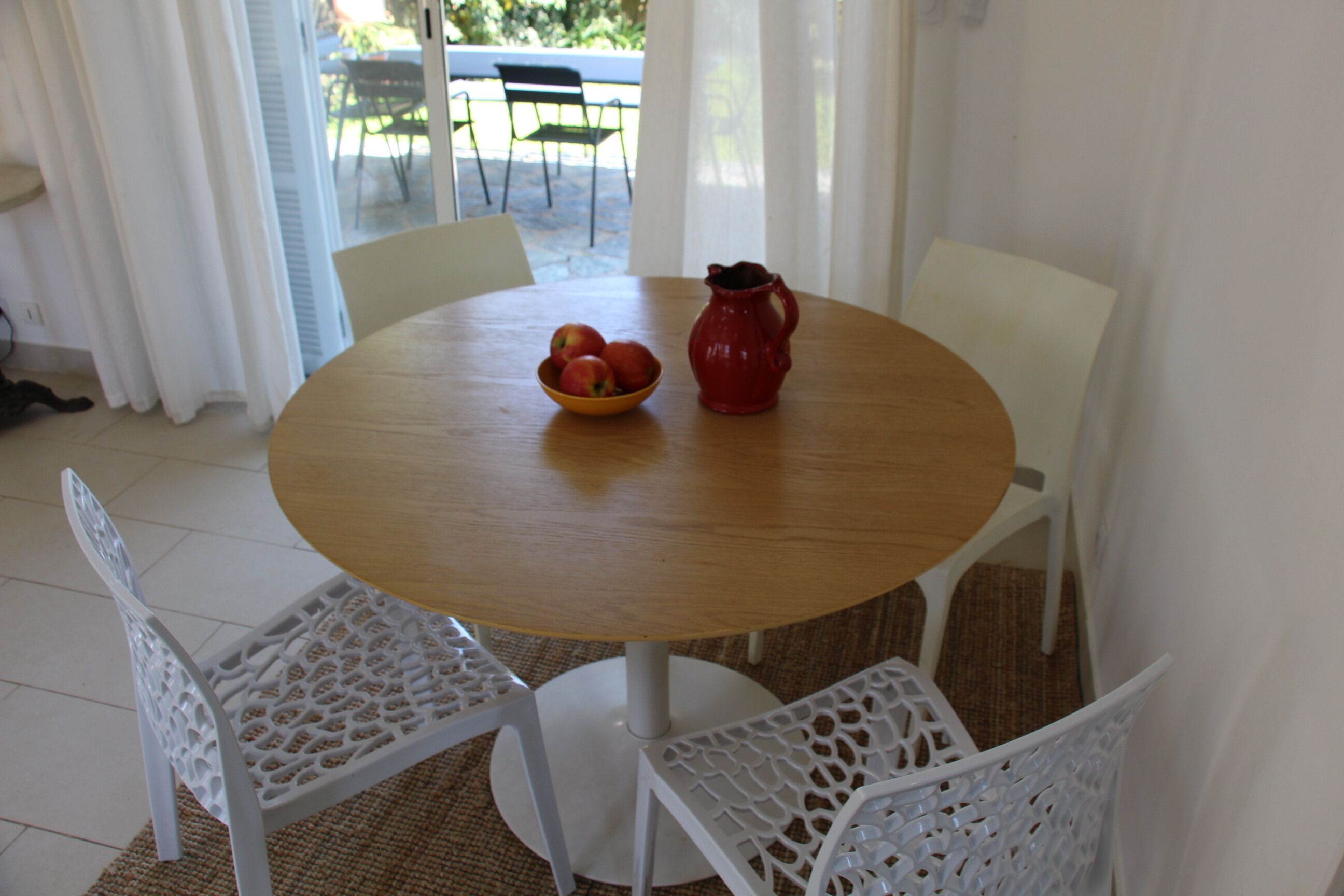 A round wooden table with a bowl of fruit and a red pitcher. There are four white chairs around the table.
