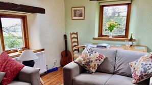 A cozy living room with a grey sofa, a red rug, a wooden guitar, and a dining table with a vase of flowers. - Detached, romantic, homely barn. Peaceful with lovely views. Garden. New kitchen (Kendal)
