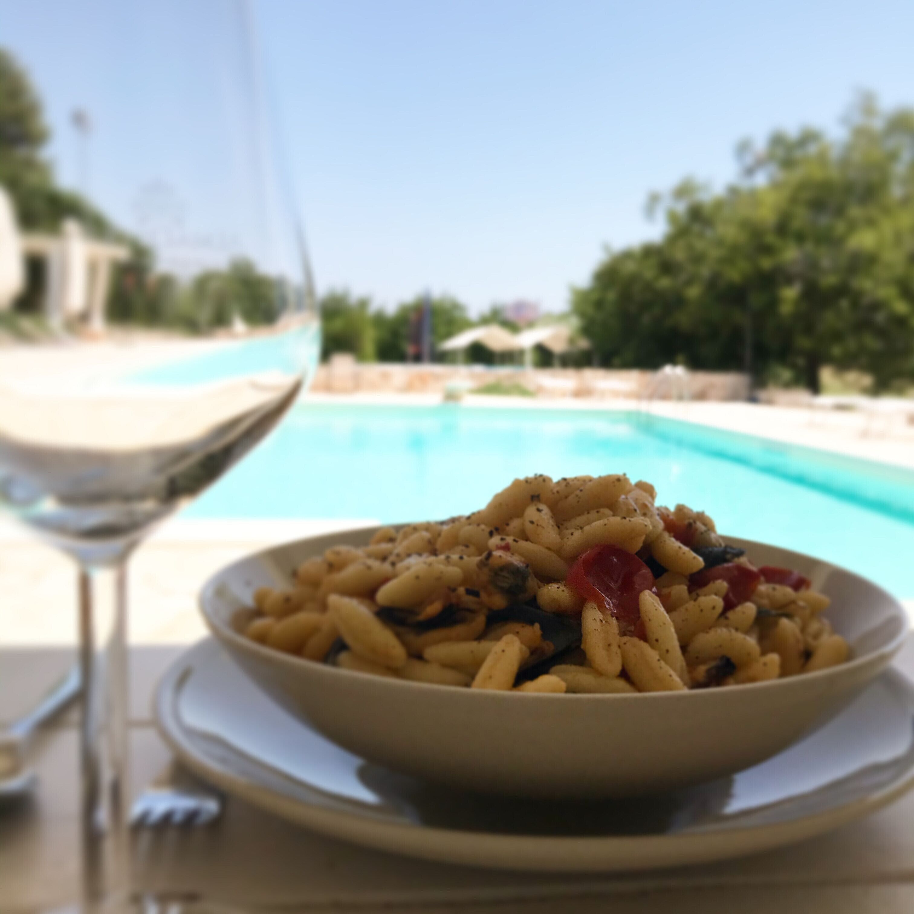 A bowl of pasta with a glass of wine on a table by a pool.