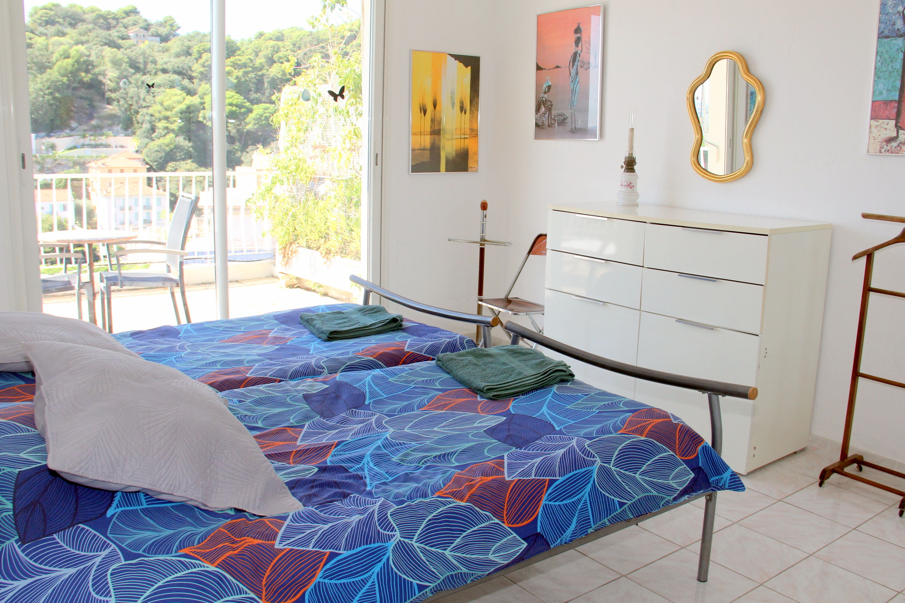 A bedroom with a bed, a dresser, a chair, and a balcony with a view of trees and buildings.