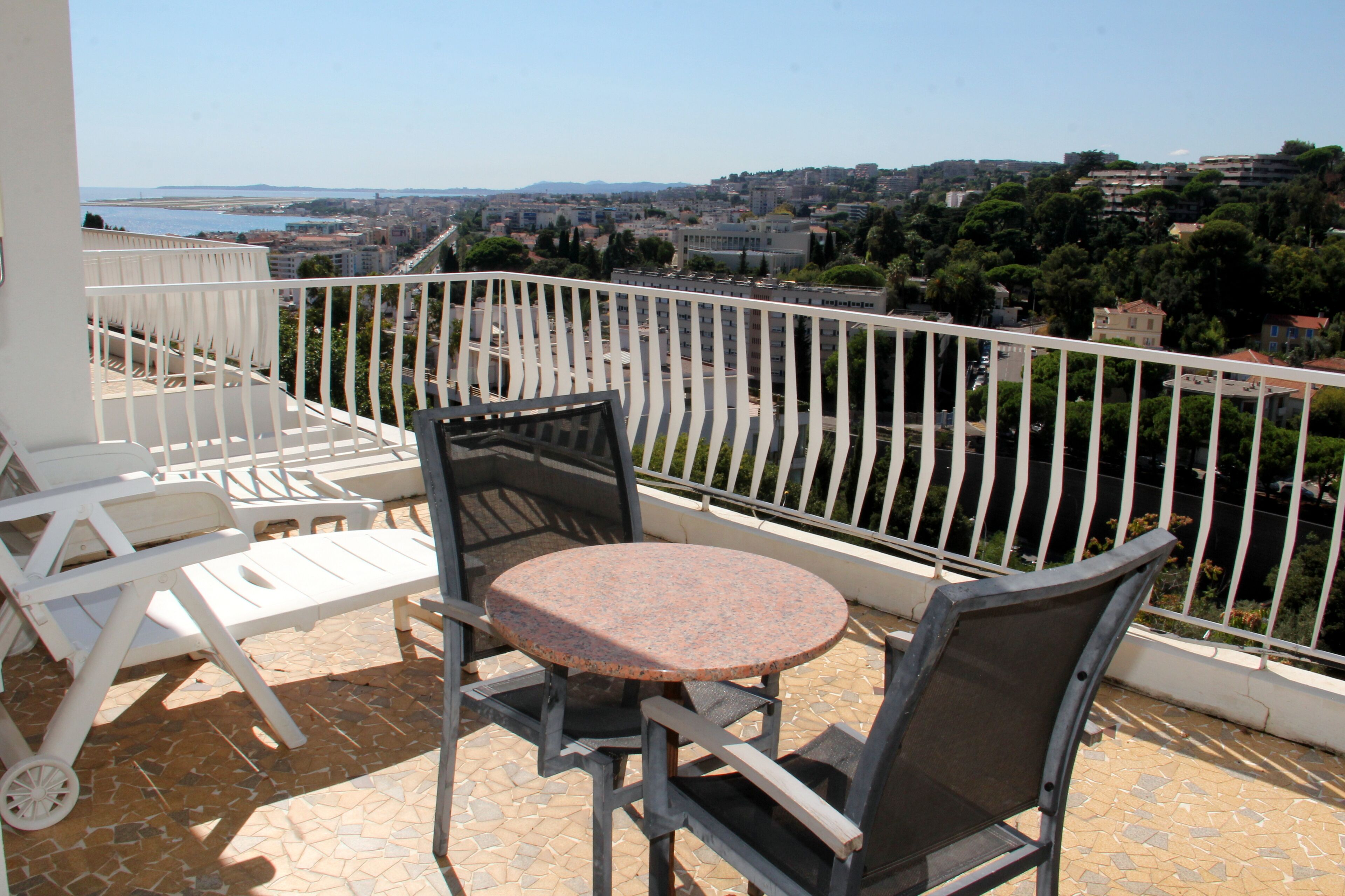 Un balcon avec une table et deux chaises, donnant sur un paysage urbain avec des bâtiments et de la verdure.