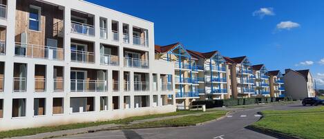 A modern multi-story residential building with balconies, a parking area, and a clear sky.