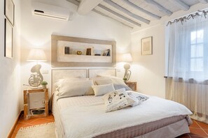 Ein Schlafzimmer mit einem Bett, zwei Nachttischen, einer Wandregal mit Büchern und Dekoration sowie einem Fenster mit durchsichtigen Vorhängen und einem Deckenventilator.