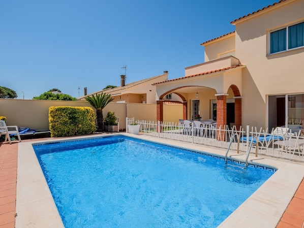 A swimming pool with a white fence, lounge chairs, and a building with a tiled roof and arches.