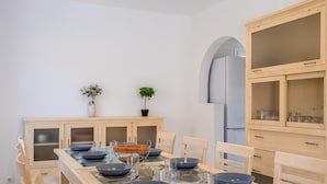 Un comedor con una mesa de vidrio, platos azules y sillas de madera. Hay dos plantas en macetas en la pared y un aparador con puertas de vidrio a la derecha.