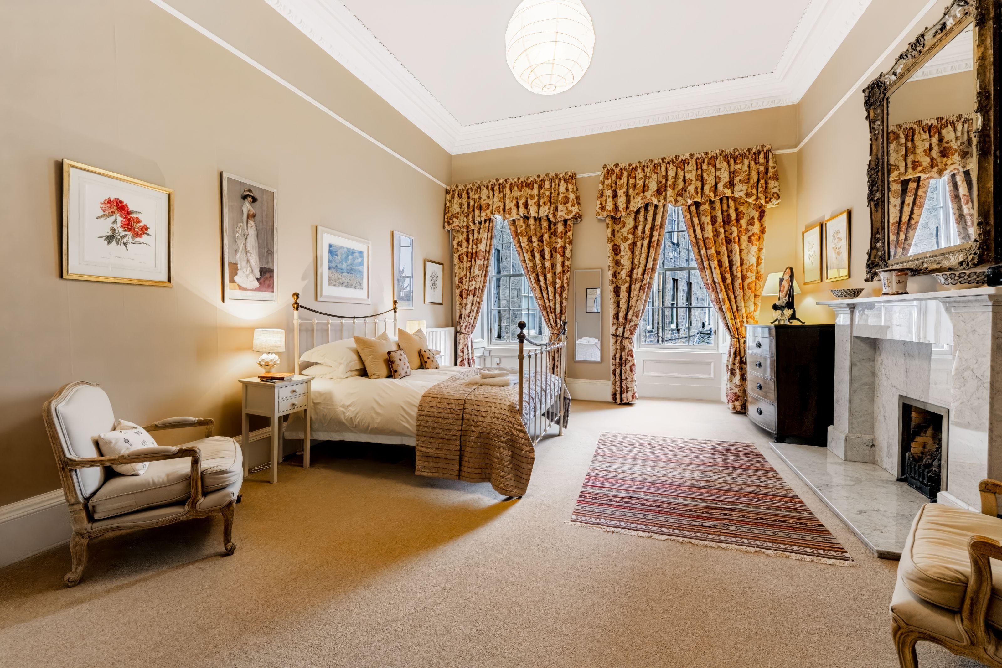 A well-appointed bedroom with a large bed, a sitting area with a chair, a fireplace, and artwork on the walls.