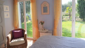 A bedroom with a bed, a wicker chair with a cushion, a small table with a vase, and a window with curtains. - Beautiful house with big garden and private pool (Mazan)