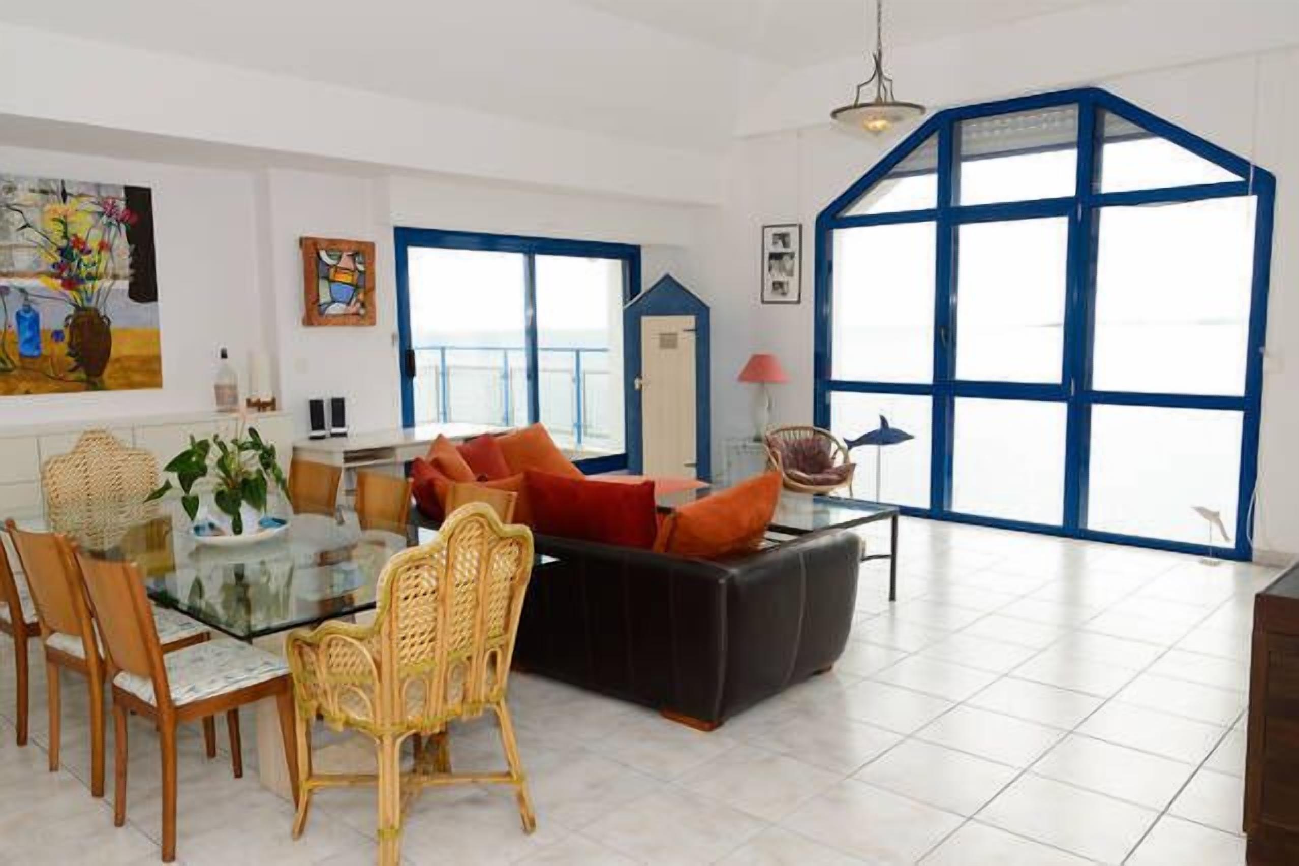 Un salón amplio con una mesa de comedor de vidrio, asientos cómodos y ventanas grandes con marcos azules.