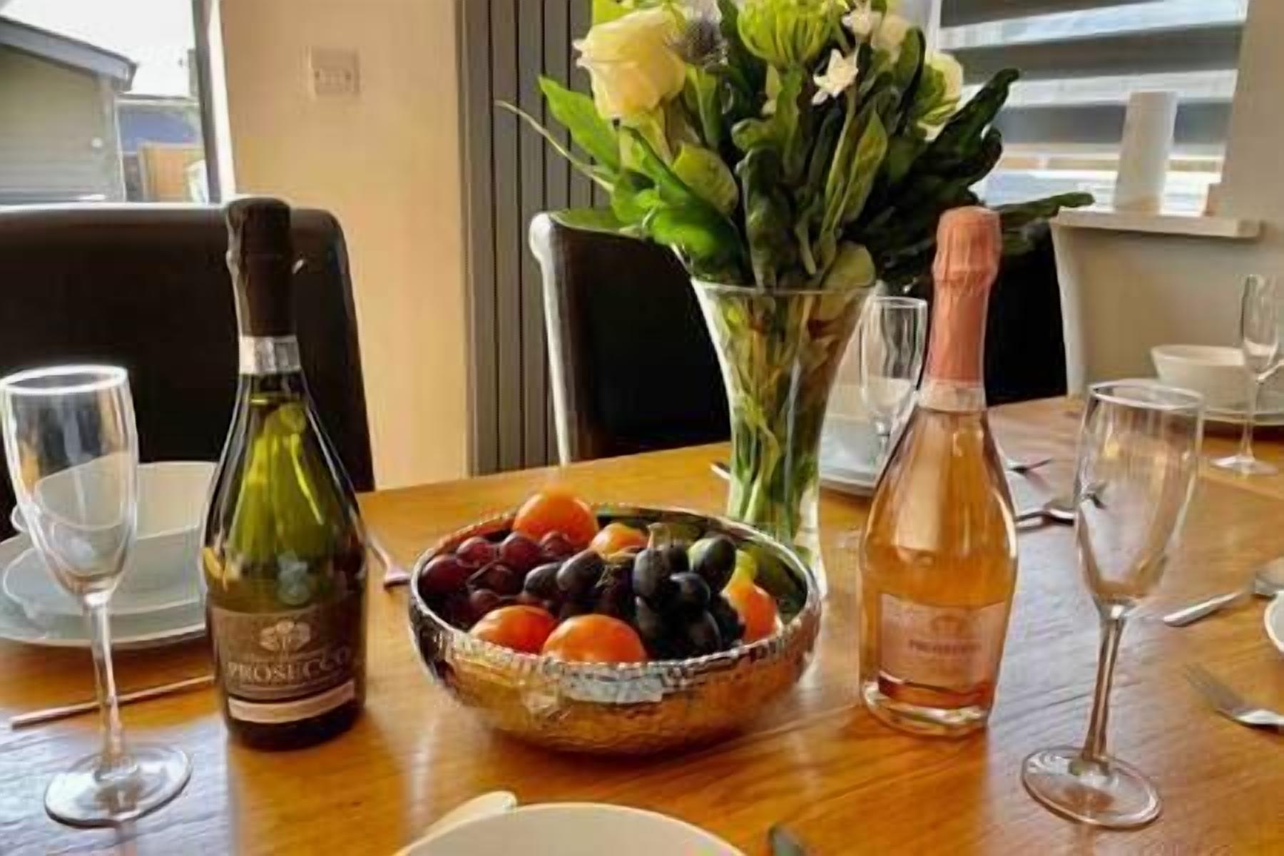 A dining table with a bottle of wine, a bowl of fruit, and a vase with flowers.