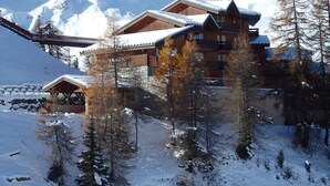 Una cabaña de madera con varios balcones y fachada de piedra, rodeada de árboles y montañas cubiertas de nieve.