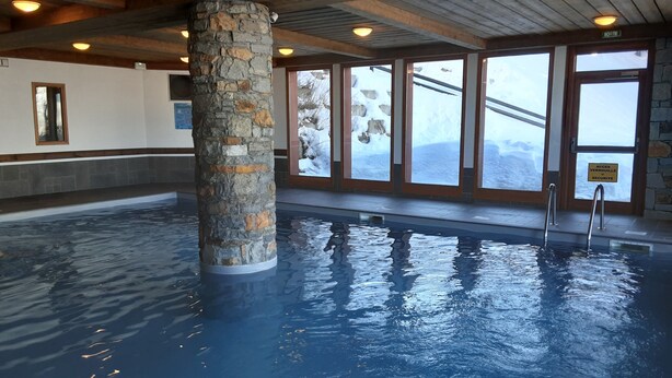 Una piscina interior con pilares de piedra, amplias ventanas y vistas a montañas nevadas.