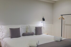 A neatly made bed with white and gray linens, a bedside table with a lamp, and a wooden hanger on a wall-mounted rack.