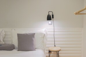 Ein ordentlich bezogenes Bett mit weißen und grauen Kissen, ein Nachttisch mit Lampe und eine Wandleuchte.