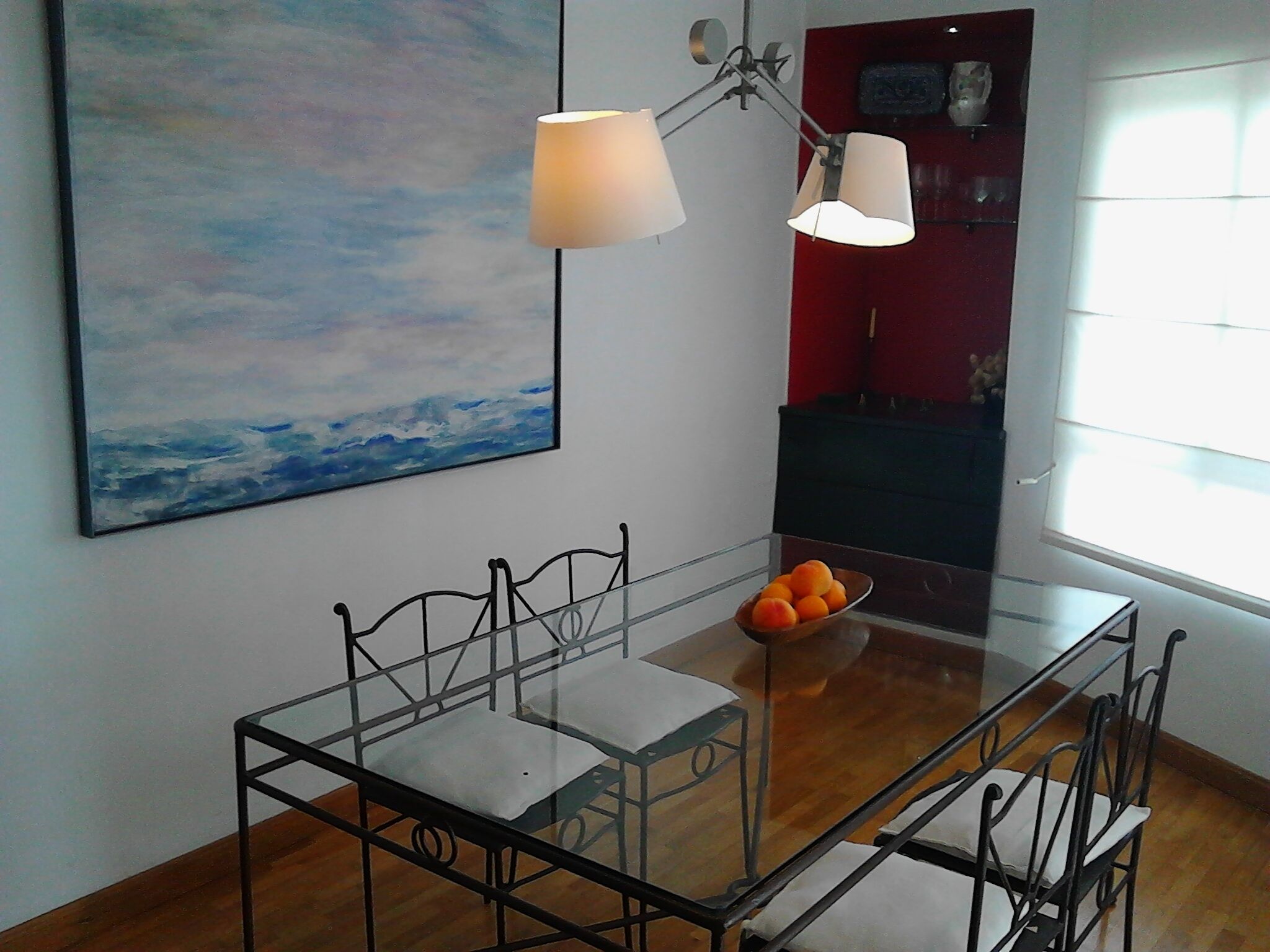 Un coin repas avec une table en verre, des chaises en métal, un saladier rempli de fruits et un tableau représentant un paysage marin accroché au mur.
