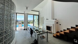 A modern living room with a glass wall, a fireplace, a dining area, and a view of a pool and mountains.