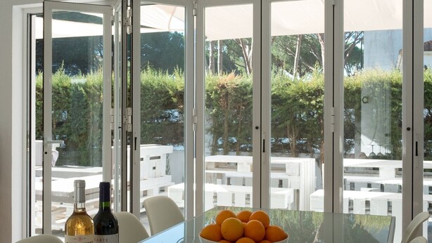 A dining area with a glass table, white chairs, a bowl of oranges, wine bottles, and a view of a pool and greenery outside.