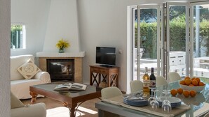 A modern living room with a fireplace, a television, a dining table with chairs, and a view of the outdoors through large windows.