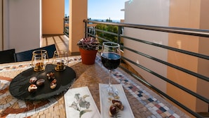 Un balcon avec une table dressée pour deux personnes, comprenant une bouteille de vin rouge, deux verres de bière et une assiette de noix.