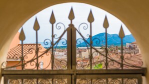 A view through an arched window with a metal railing, overlooking a town with terracotta rooftops and a mountain in the background.