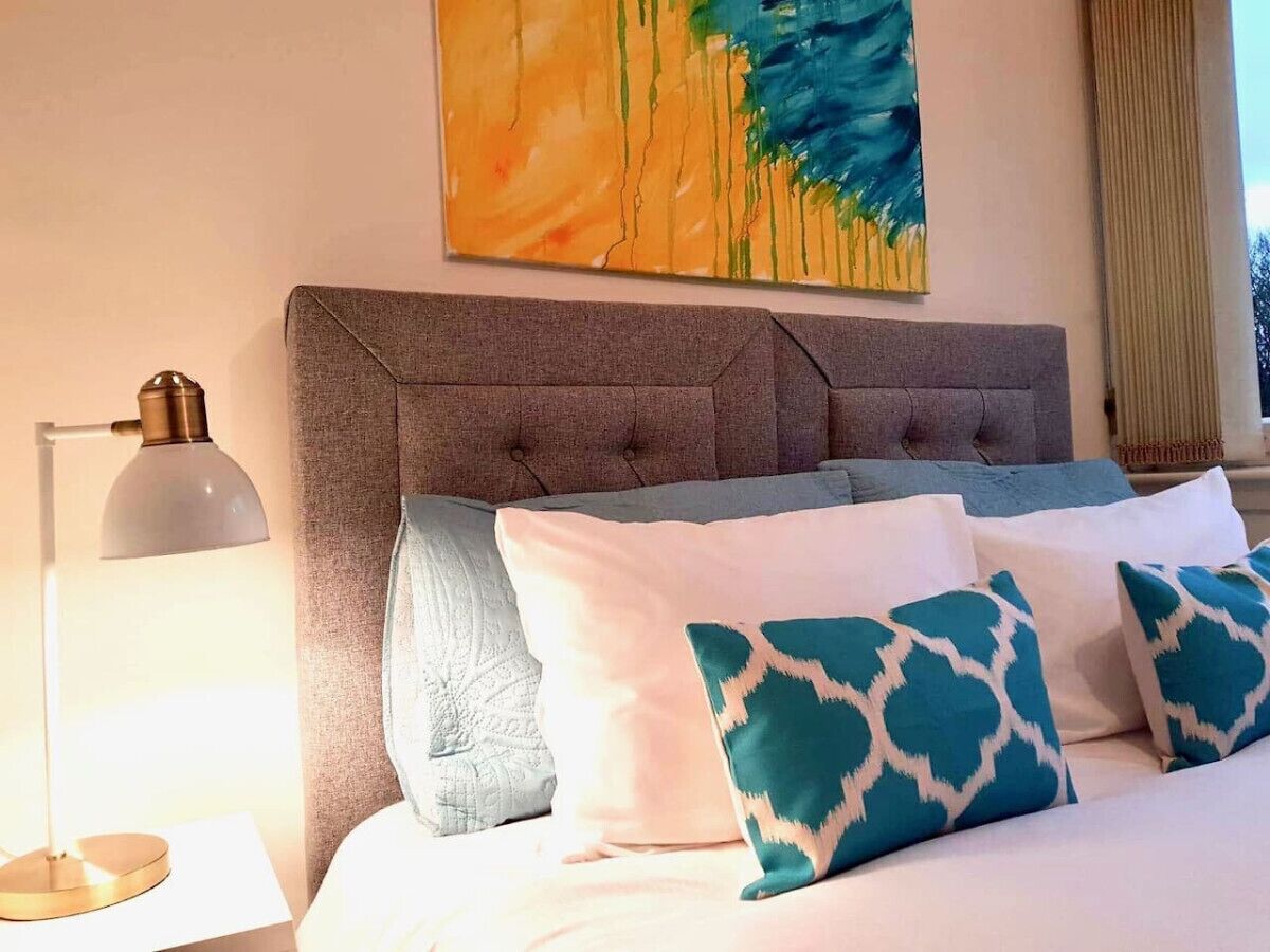 A neatly made bed with patterned pillows, a grey upholstered headboard, and a modern lamp on a bedside table.