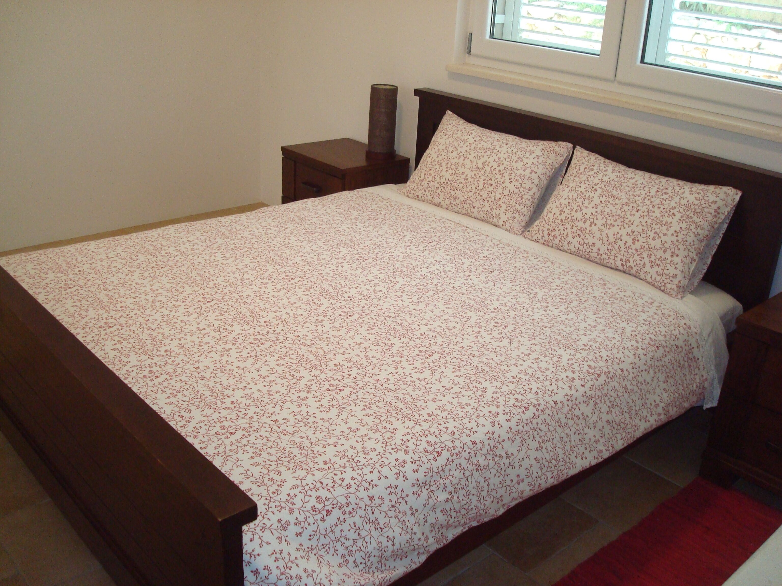 Una cama con un edredón estampado con motivos florales y dos almohadas, una mesita de noche de madera con una lámpara, y una ventana con persianas.