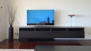 A flat-screen TV mounted on a dark wooden entertainment center with a glass top, a vase with branches, and a clear glass table with a red object on it. - Excellent beach front apartment with large sunny balcony overlooking the sea (Vila Nova de Gaia)