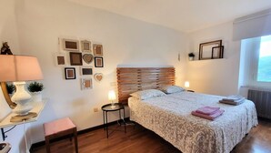 Un dormitorio con cabecera de madera, cama con ropa de cama estampada, mesita de noche con lámpara y un pequeño taburete.