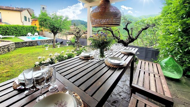Un área de comedor al aire libre con mesas y sillas de madera, un farol colgante y una vista a una piscina y jardín al fondo.