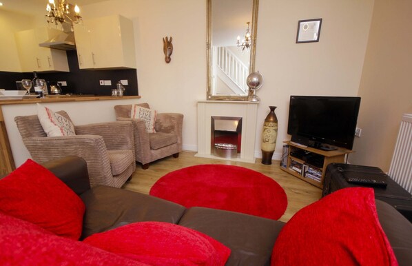 A living room with a fireplace, a television, and a kitchen area in the background.