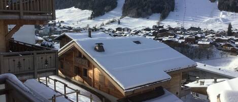 Un chalet en bois, dont le toit est recouvert de neige, avec un balcon doté d’une balustrade, et qui offre une vue sur un village enneigé et des montagnes en arrière-plan.